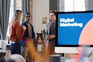 Best Freelance Digital Marketer in Dubai,three digital marketing professionals in a modern office discussing strategy in front of a large monitor displaying 'Digital Marketing' text, with a blurred foreground of desk items.